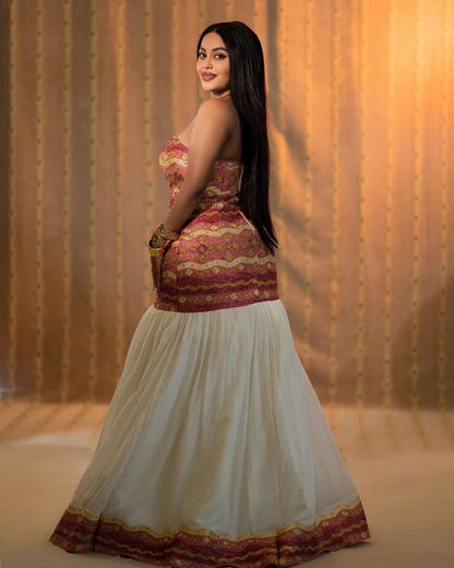 Woman wearing a traditional Ethiopian dress with a patterned top and white skirt against a warm-toned curtain background.