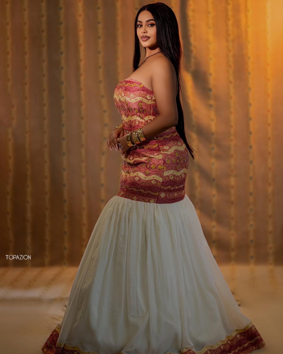 Woman wearing a strapless top with red and gold Ethiopian dress and a white skirt against a textured wall.