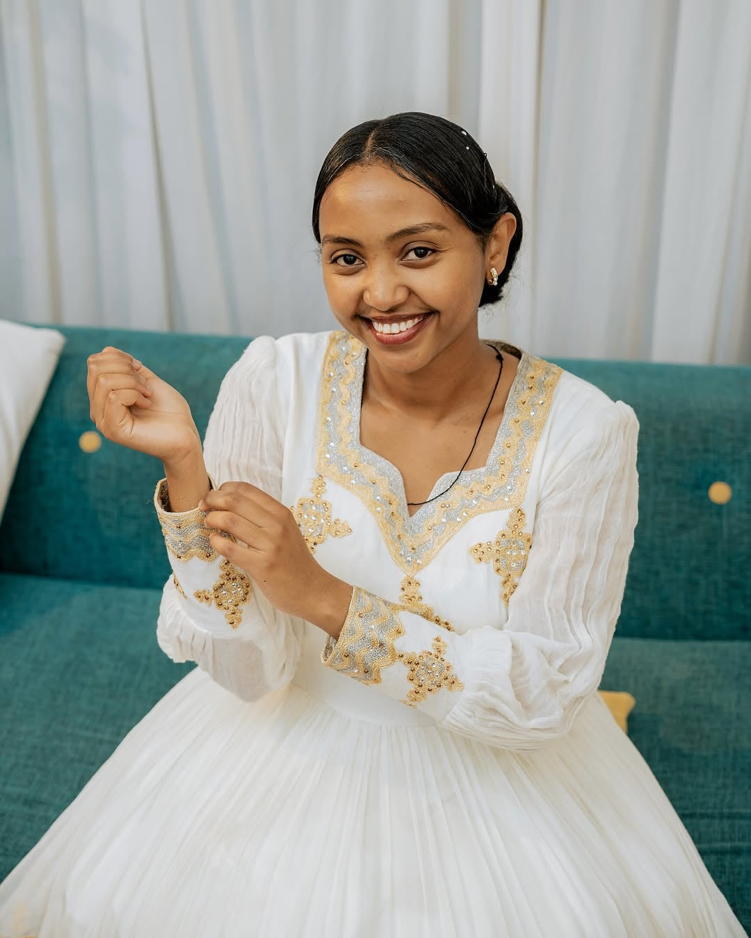 Woman in a white dress with gold embroidery sitting on a teal couch.