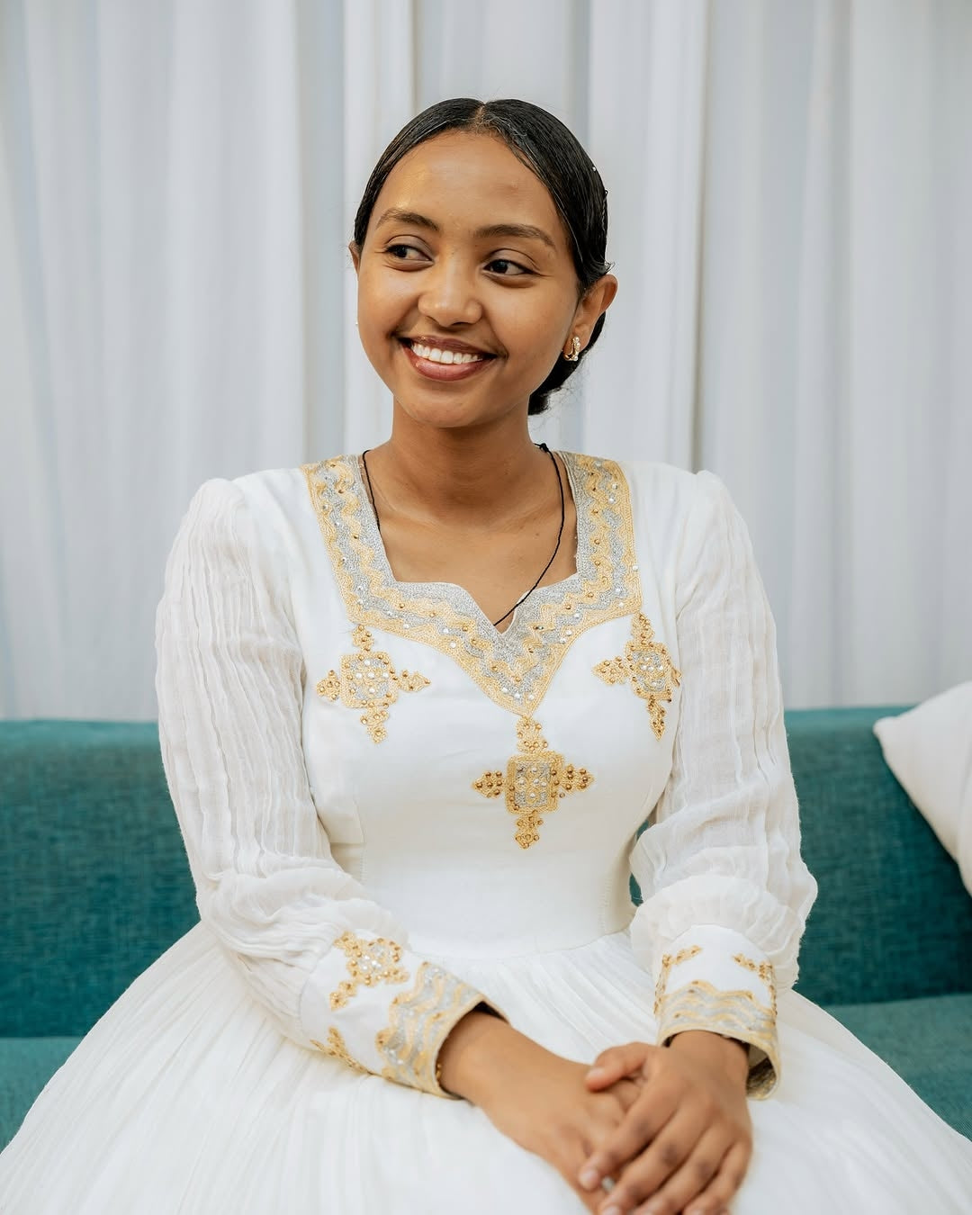 Woman in a white dress with gold embroidery sitting on a teal couch against a white curtain background.