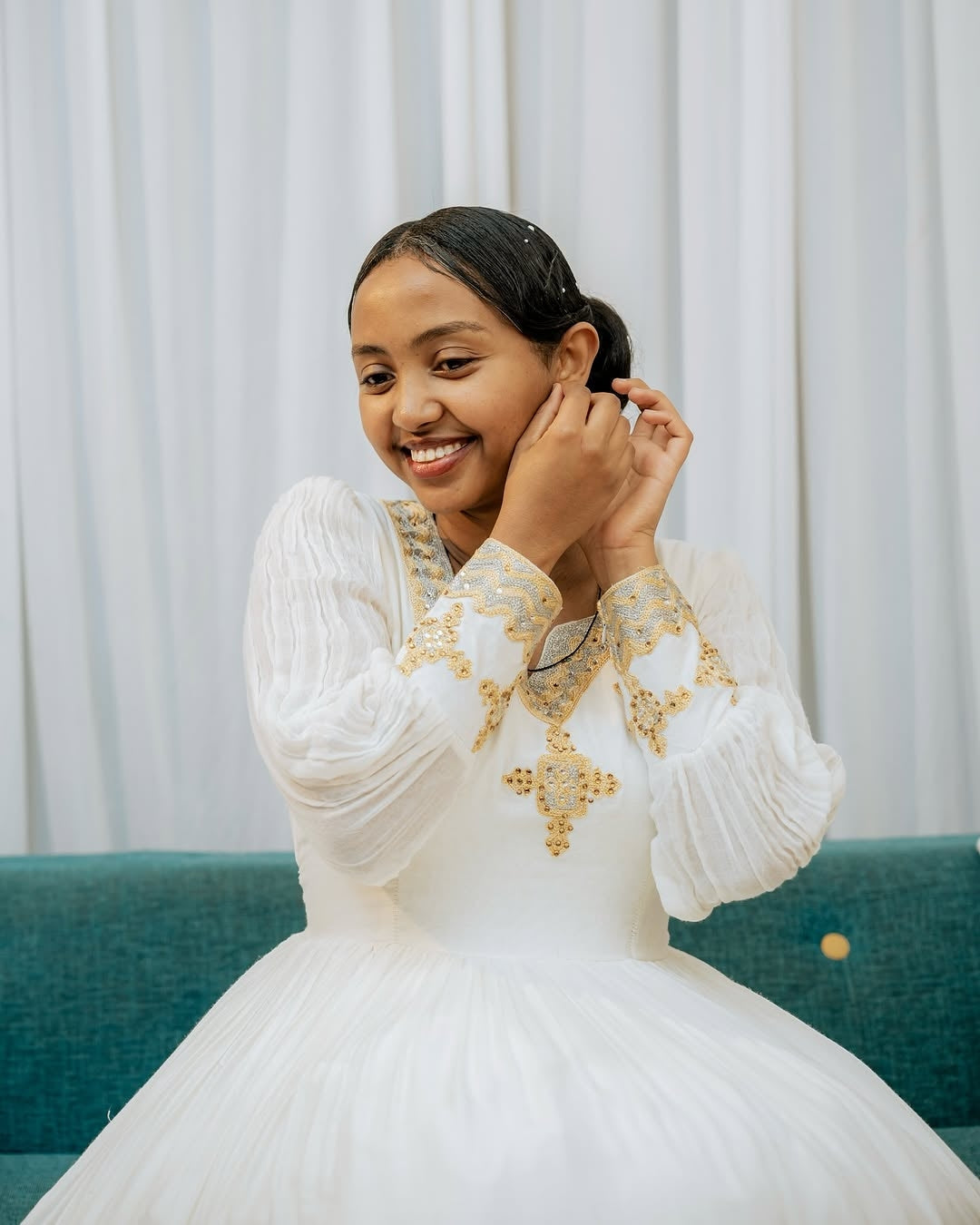 Woman in a white dress with gold embroidery sitting on a teal couch against a white curtain background