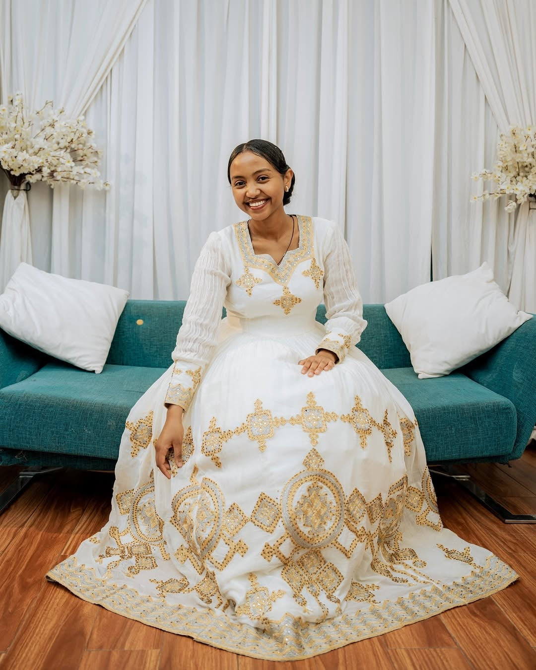 Woman in a white and gold embroidered dress sitting on a teal couch with a white curtain background.