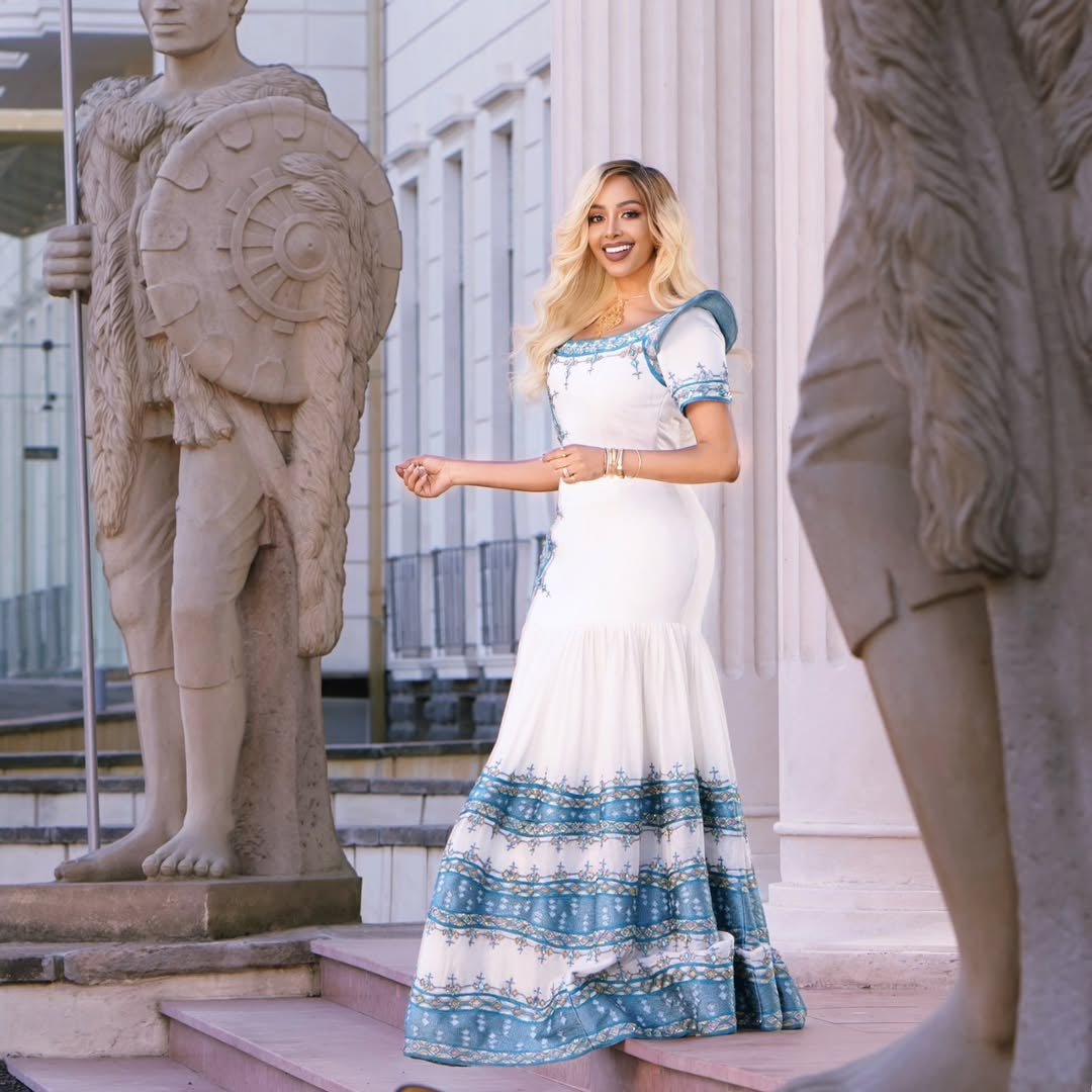 Woman in a white and blue habesha kemis standing between two stone statues in an outdoor setting.