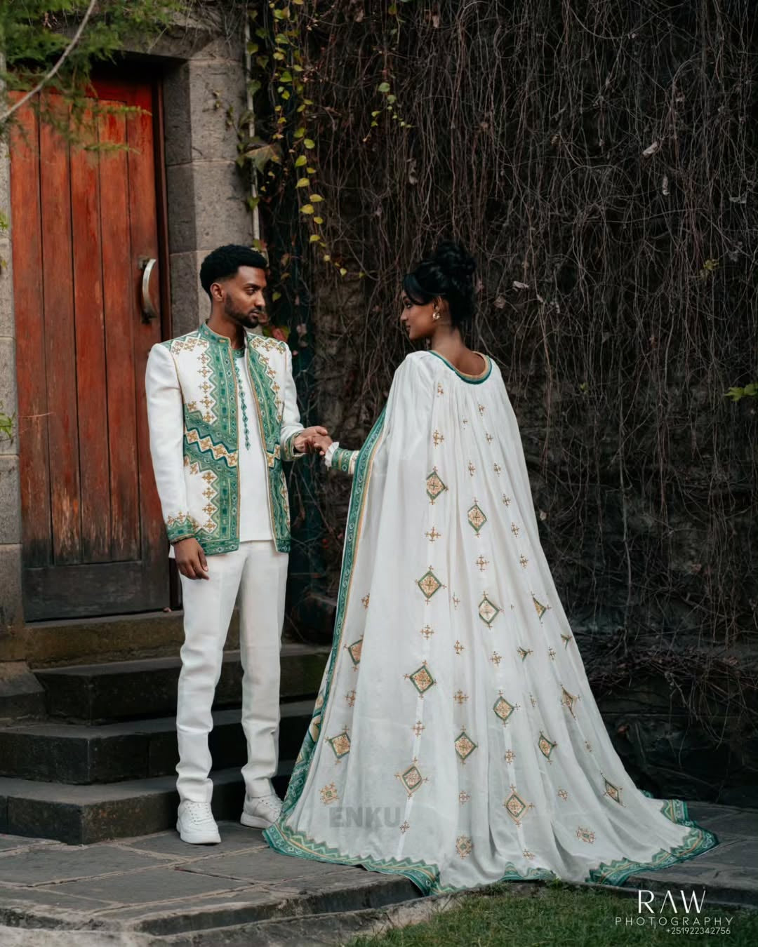 Man and woman in traditional Habesha Outfit couple Set  standing outdoors with a wooden door and ivy background.