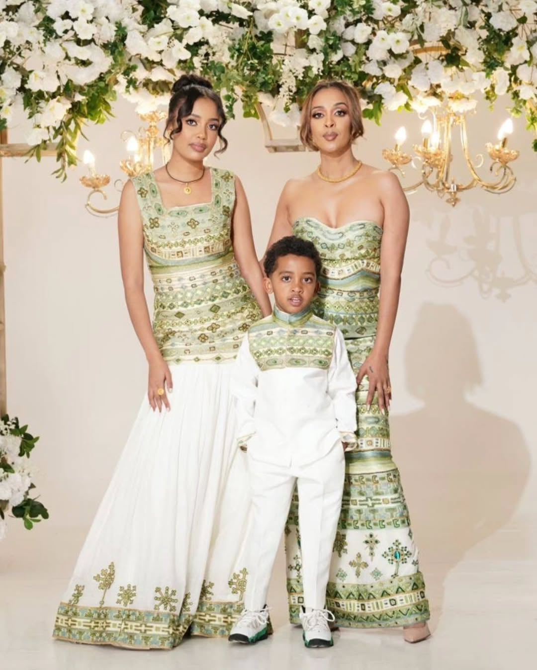 Three people in matching green and white habesha kemis  standing in front of floral decorations.