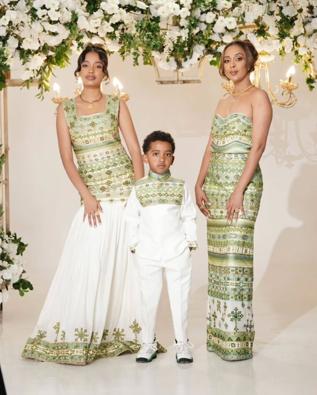 Two women and a child in matching green and white Habesha Dress  standing in front of floral decorations.