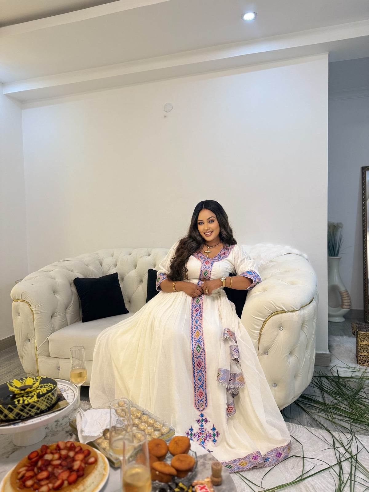 A woman wearing a traditional white embroidered Ethiopia clothes with colorful blue, red, and purple patterns sits on a tufted cream sofa in a modern living room. A table in the foreground holds food and drinks.