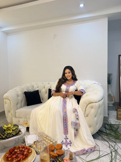 A woman wearing a traditional white embroidered Ethiopia clothes with colorful blue, red, and purple patterns sits on a tufted cream sofa in a modern living room. A table in the foreground holds food and drinks.
