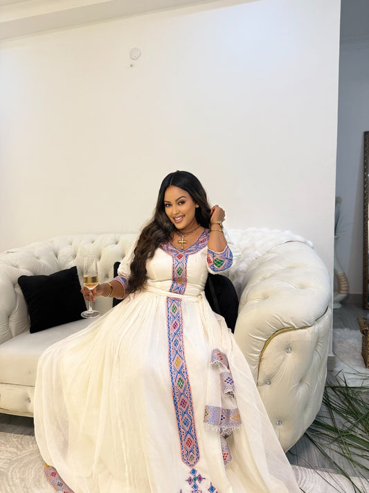A woman wearing a white traditional Ethiopia clothes with colorful embroidered details sits on a white tufted sofa, holding a glass of champagne in a modern living room setting.