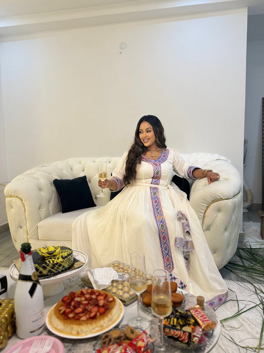 A woman wearing a white traditional Ethiopia clothes with colorful embroidered details sits on a white tufted sofa, holding a glass of champagne in a modern living room setting.