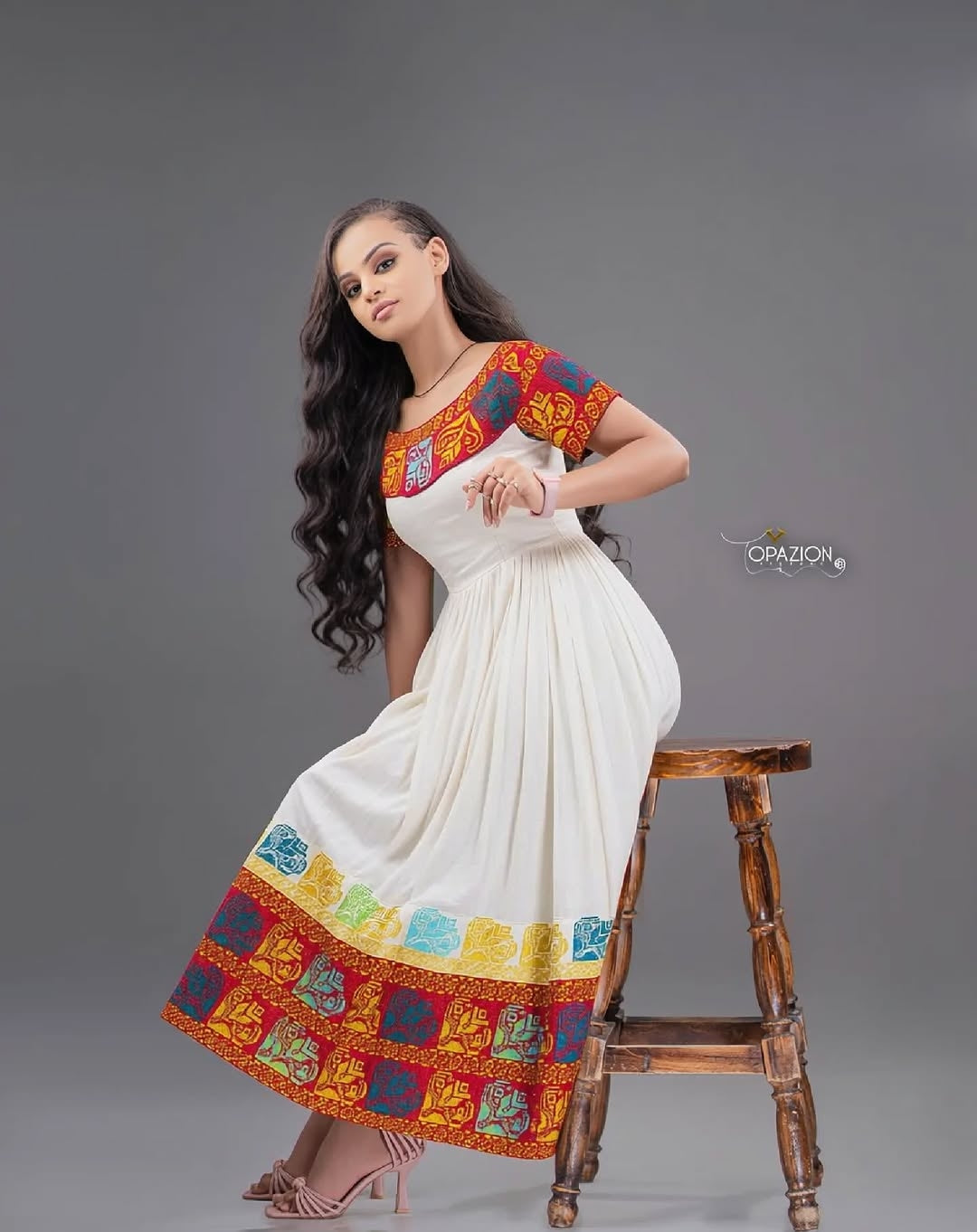 Woman wearing a colorful Red Ethiopian Traditional Dress with a white base, sitting on a wooden stool against a gray background.