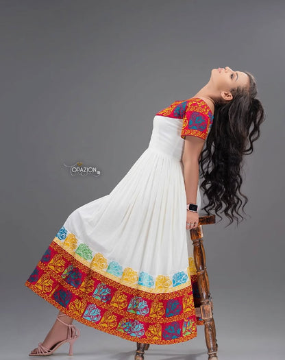 Woman wearing a white Red Ethiopian Traditional Dress with colorful embroidery, leaning on a wooden stool against a gray background.