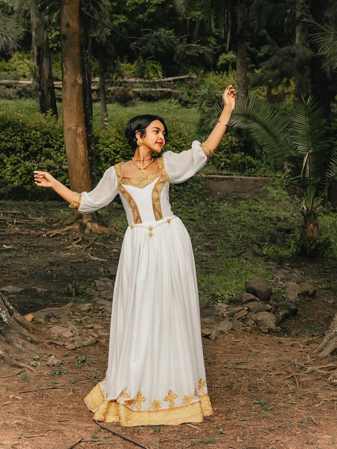 Woman in a white and gold dress standing in a natural setting with trees and greenery.