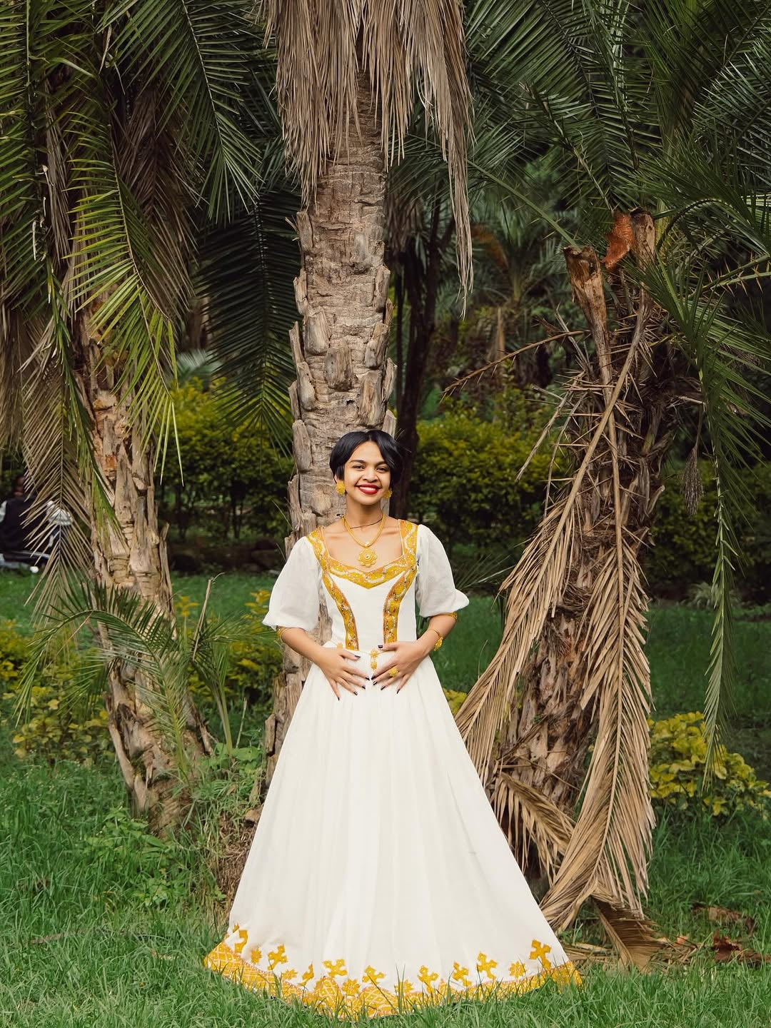 Woman in a white and yellow dress standing in a garden with palm trees.