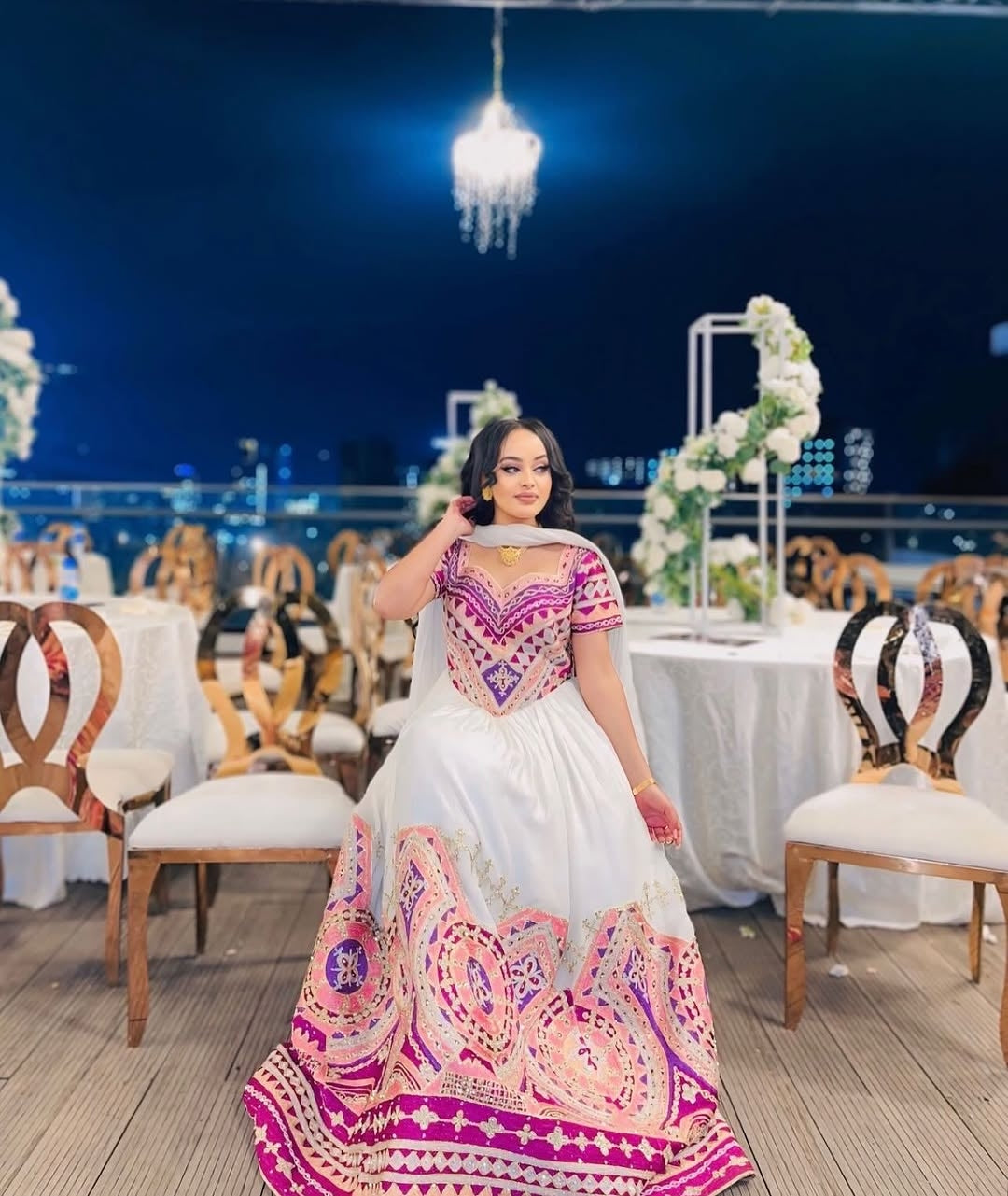 Woman in a colorful traditional habesha wedding outfit standing in a decorated indoor setting with tables and chairs.