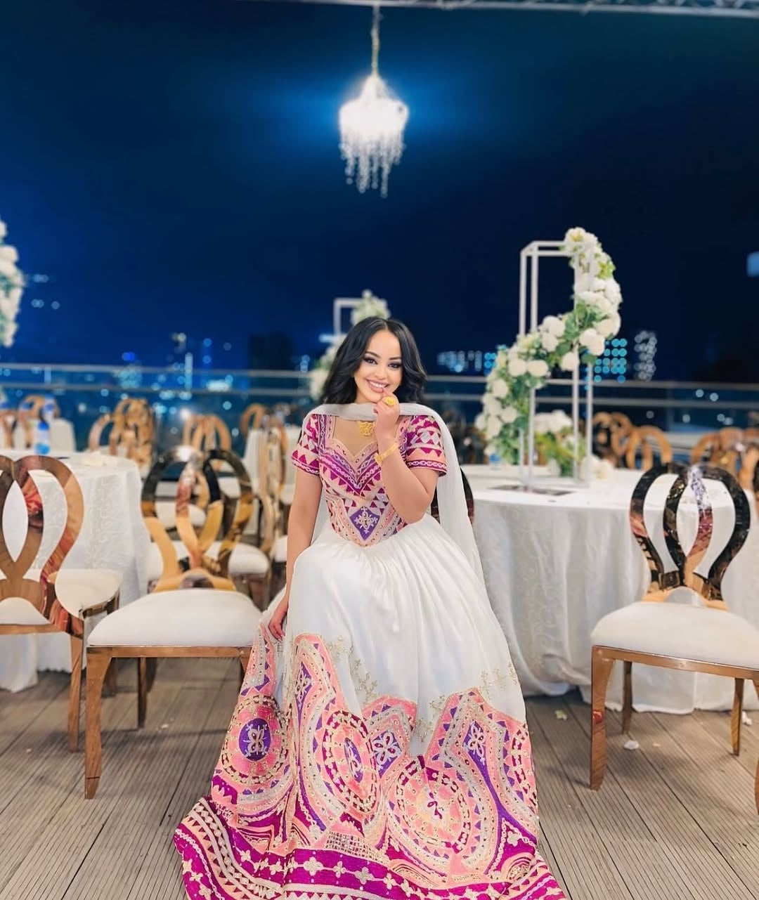 Woman in a pink habesha wedding dress standing in a decorated indoor setting with tables and chairs.