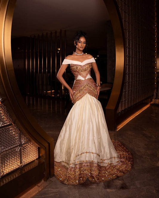 Woman in a gold and white habesha dress standing in front of a decorative mirror.