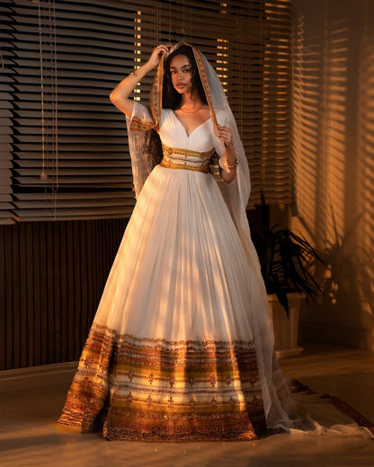 Woman in a white and gold evening Ethiopian cultural dress with a long train, standing indoors with blinds in the background.