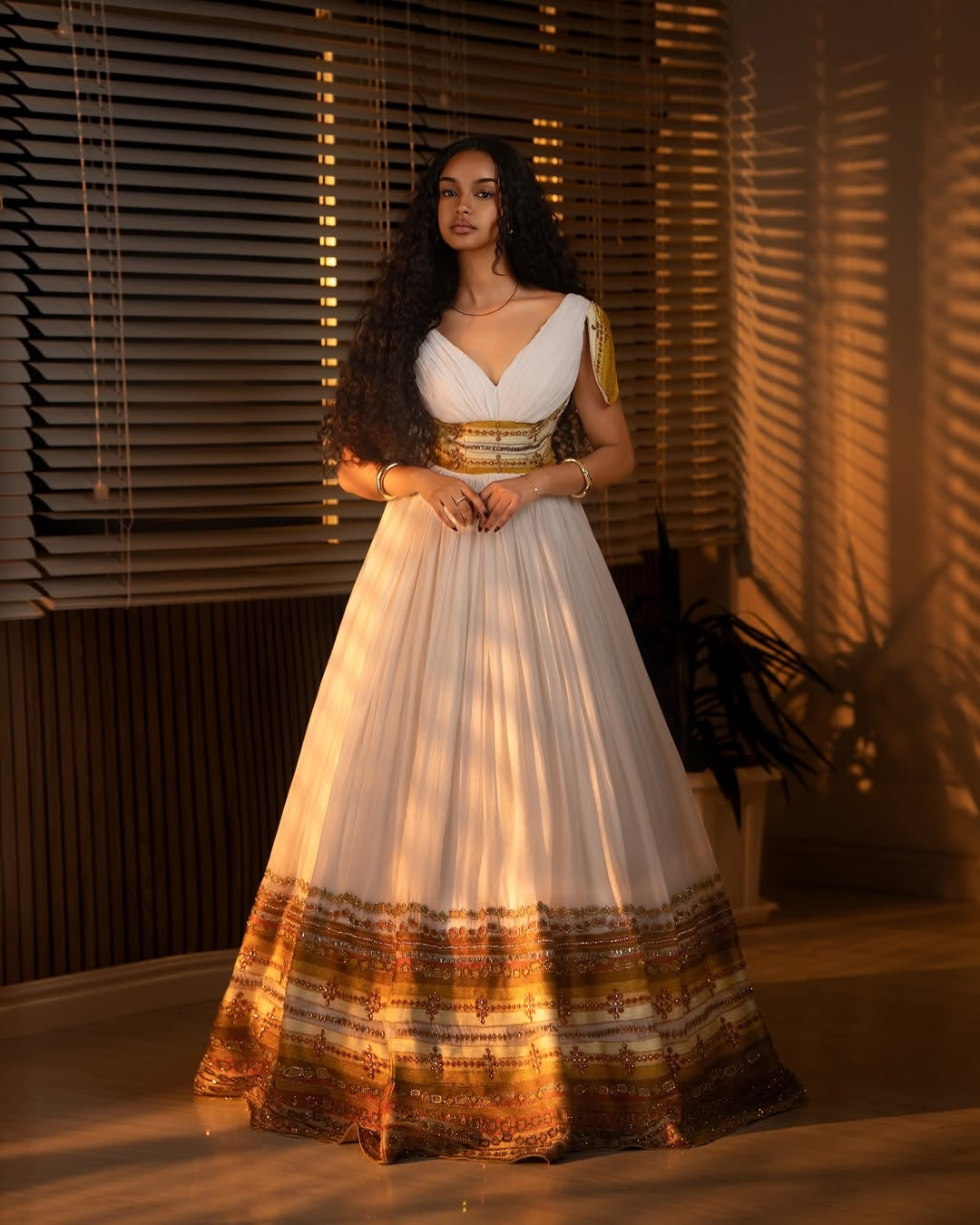 Woman in a white and gold traditional Ethiopian cultural dress standing in a warm-lit room.