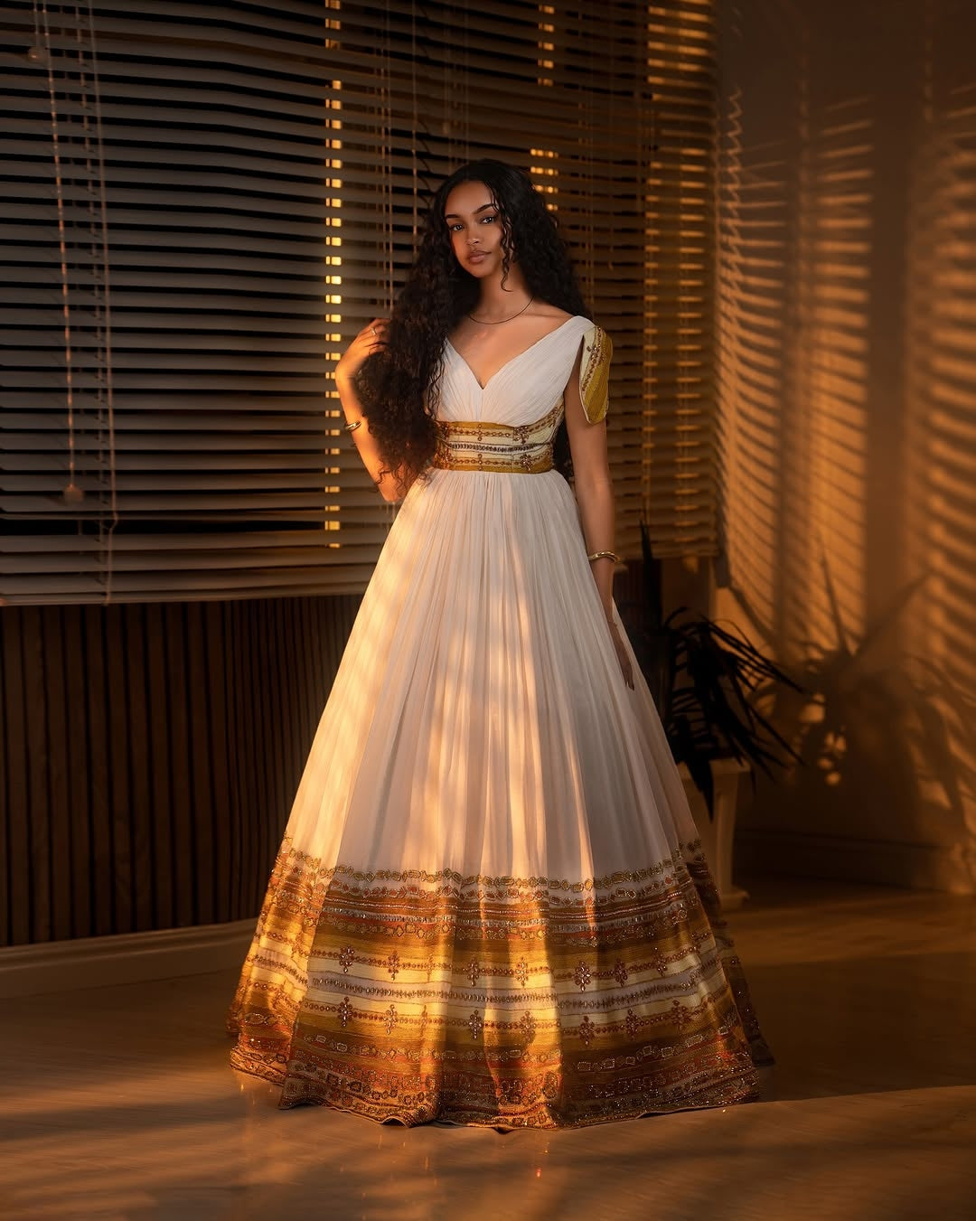 Woman in a white and gold Ethiopian cultural dress standing in a room with warm lighting.