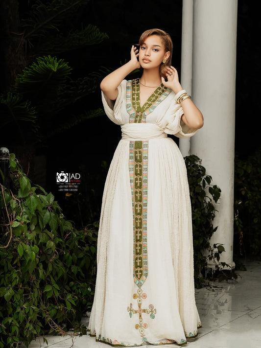 Woman wearing a long, white Ethiopian cultural dress with green embroidery in an outdoor setting.