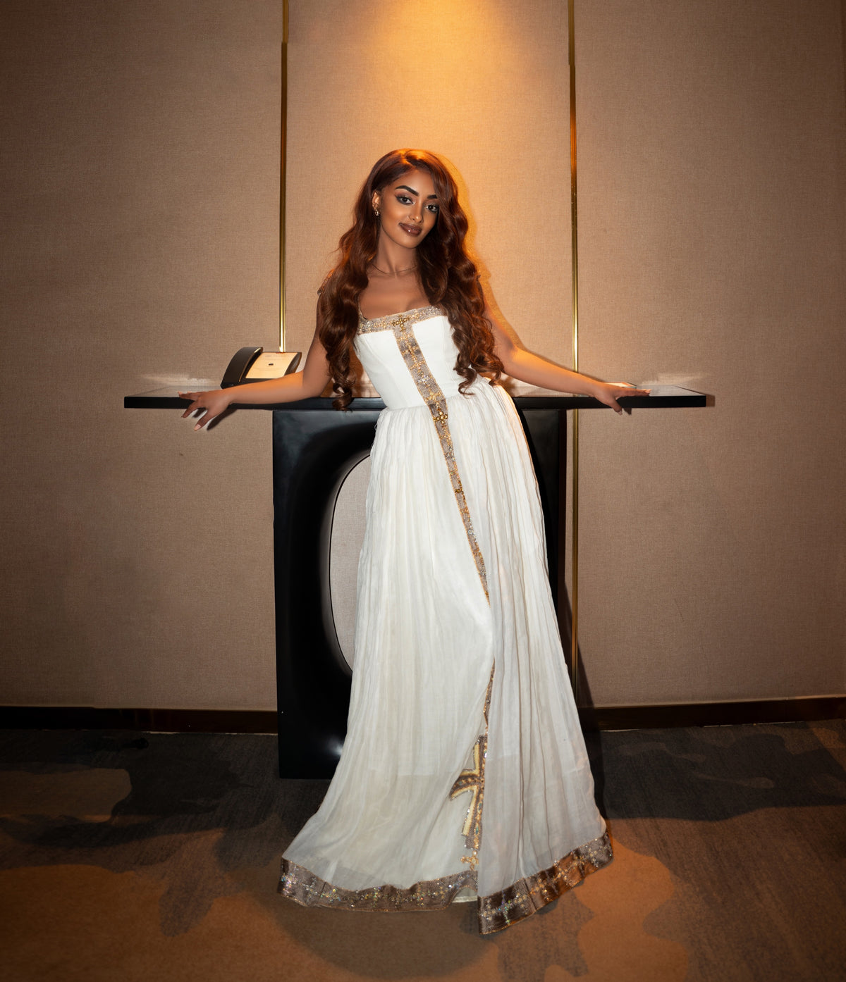A woman wearing a strapless white floor-length Ethiopian clothing with gold embroidered trim stands behind a black display table in an indoor setting with warm wall lighting.