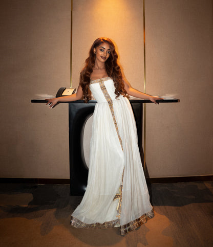 A woman wearing a strapless white floor-length Ethiopian clothing with gold embroidered trim stands behind a black display table in an indoor setting with warm wall lighting.
