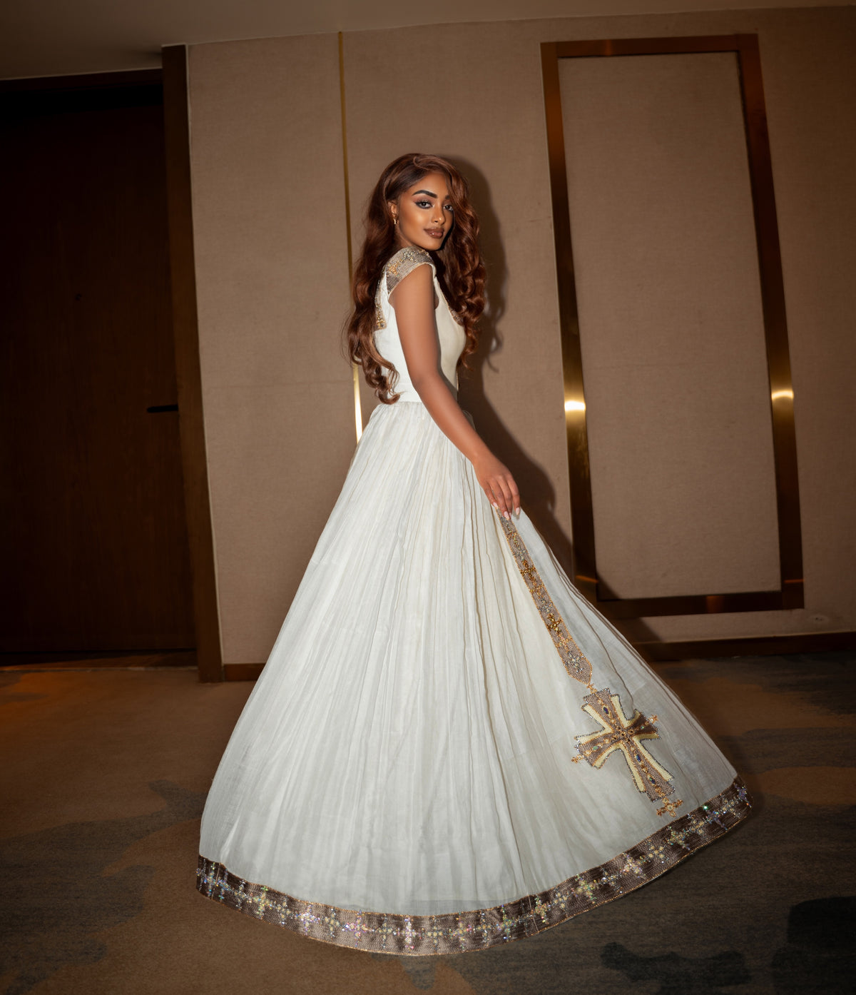 A woman wearing a white sleeveless Ethiopian clothing with gold embroidered detailing and a large flared skirt, photographed indoors against a beige wall with gold-framed panels.