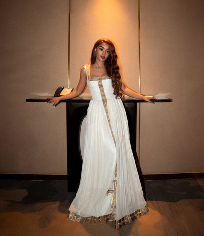 A woman wearing a long white Ethiopian clothing with gold embroidery and a square neckline, posing with her arms outstretched behind a black table in an indoor setting with warm lighting.