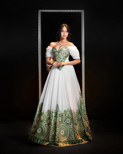 A woman stands in front of a black studio background wearing a dramatic off-the-shoulder ball gown with a white Ethiopian Traditional Clothing, green embroidered bodice, and ornate green and gold circular patterns concentrated along the hem. She is framed by a tall rectangular mirror-like border in the background.