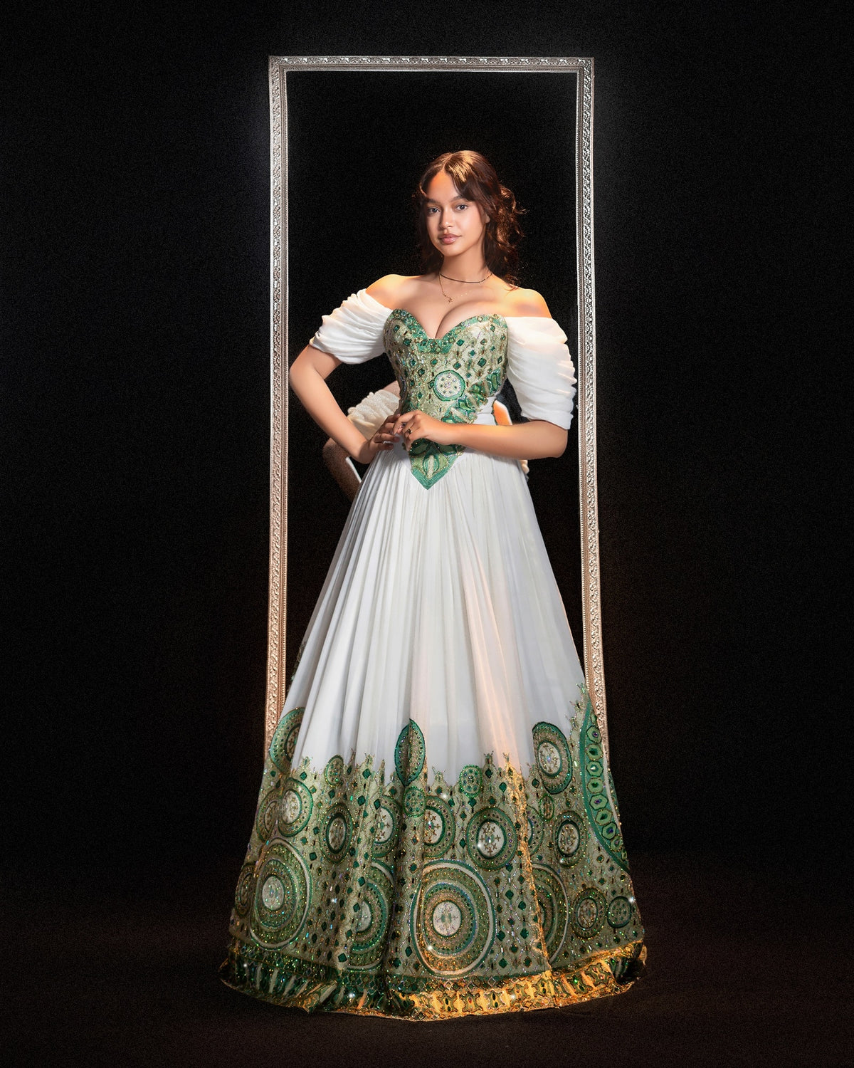 A woman stands in front of a black background wearing a floor-length off-the-shoulder Ethiopian Traditional Clothing with a white skirt, green embroidered bodice, and green-and-gold ornate border detailing along the hem. She is framed by a tall vertical mirror-like panel.