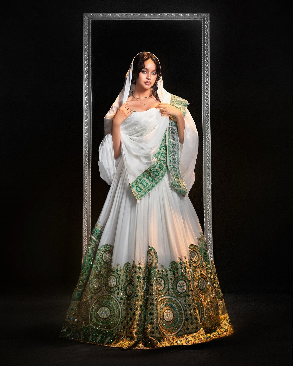 A woman stands in front of a black background wearing a white  Ethiopian Traditional Clothing with extensive green and gold circular embroidery concentrated along the hem and a matching white dupatta draped over her shoulders.