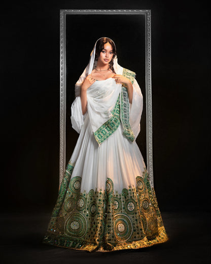 A woman stands in front of a black background wearing a white  Ethiopian Traditional Clothing with extensive green and gold circular embroidery concentrated along the hem and a matching white dupatta draped over her shoulders.