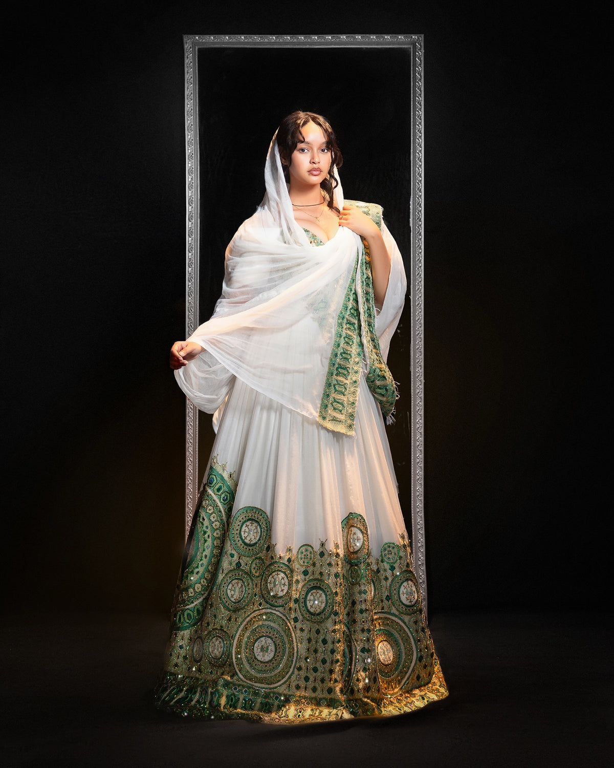 A woman models a traditional white lehenga Ethiopian Traditional Clothing with intricate green and gold embroidery, standing in front of a large decorative mirror against a black background.