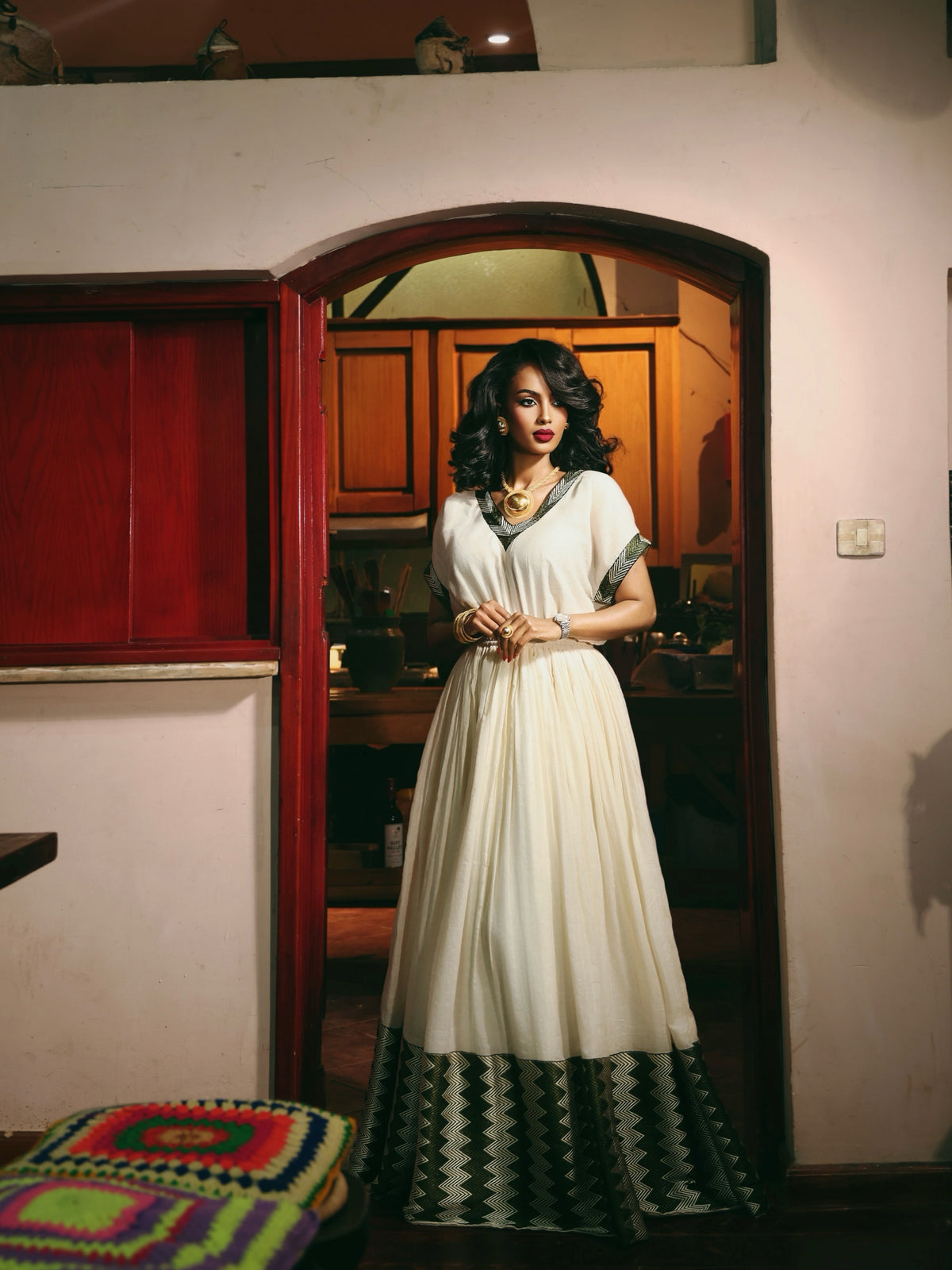 A woman stands in a doorway wearing a long cream-colored Ethiopia store  with green and black geometric patterned trim at the hem and sleeves. The setting appears to be an indoor home environment with warm lighting.
