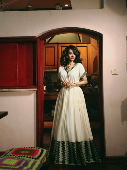 A woman stands in a doorway wearing a long cream-colored Ethiopia store  with green and black geometric patterned trim at the hem and sleeves. The setting appears to be an indoor home environment with warm lighting.