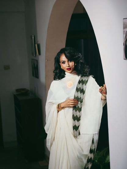 A woman wearing a flowing white Ethiopia store  with a green and white patterned shawl draped over her shoulder, standing in a doorway with a warm, dimly lit interior background.
