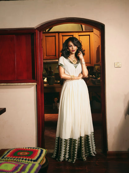 A woman stands in a doorway wearing a long white Ethiopia store  with green and black geometric patterned trim at the hem and neckline, posed in an indoor home setting with warm lighting.
