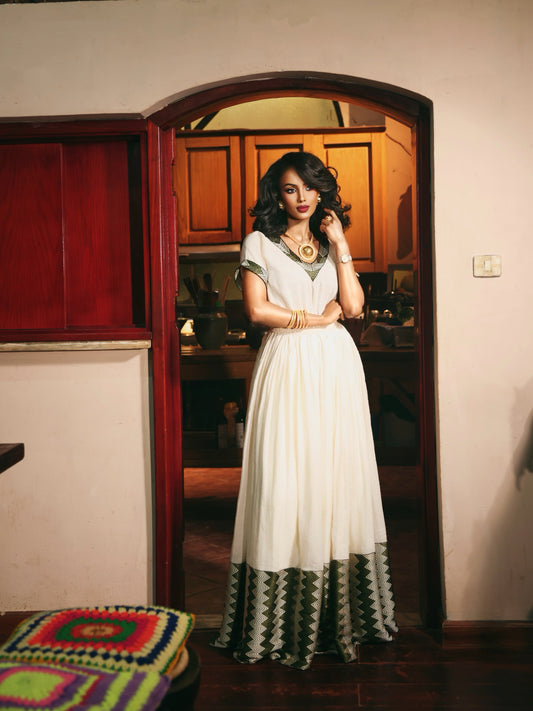 A woman stands in a doorway wearing a long white Ethiopia store  with green and black geometric patterned trim at the hem and neckline, posed in an indoor home setting with warm lighting.
