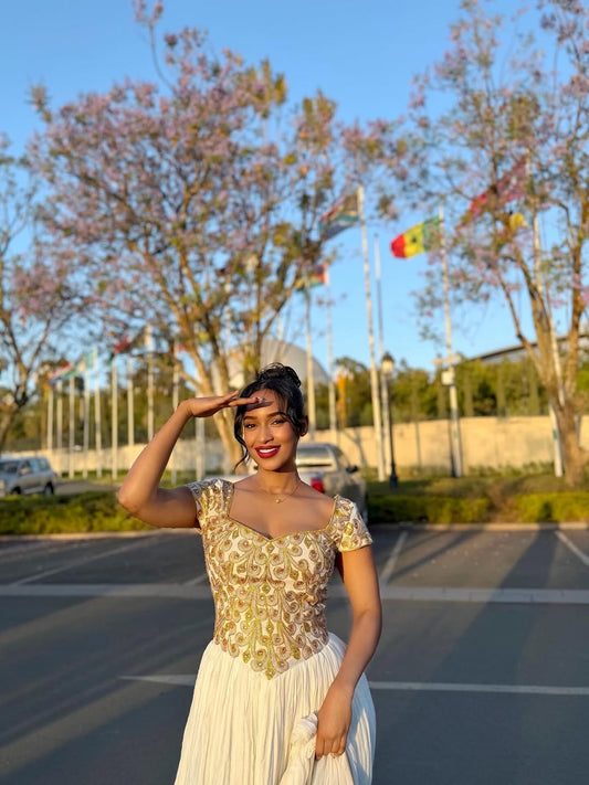 Woman in a gold and white Ethiopian cultural dress posing outdoors with trees and flags in the background
