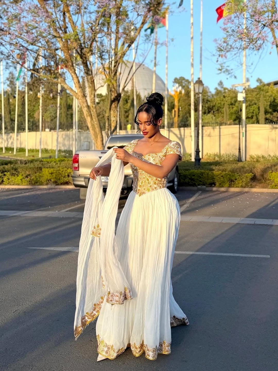 Woman in a white and gold traditional Ethiopian cultural dress standing in a parking lot with trees and flags in the background.