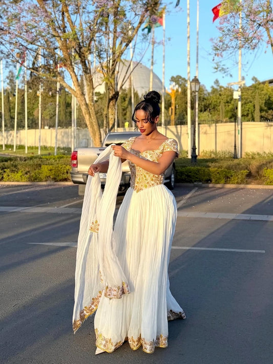 Woman in a white and gold traditional Ethiopian cultural dress standing in a parking lot with trees and flags in the background.