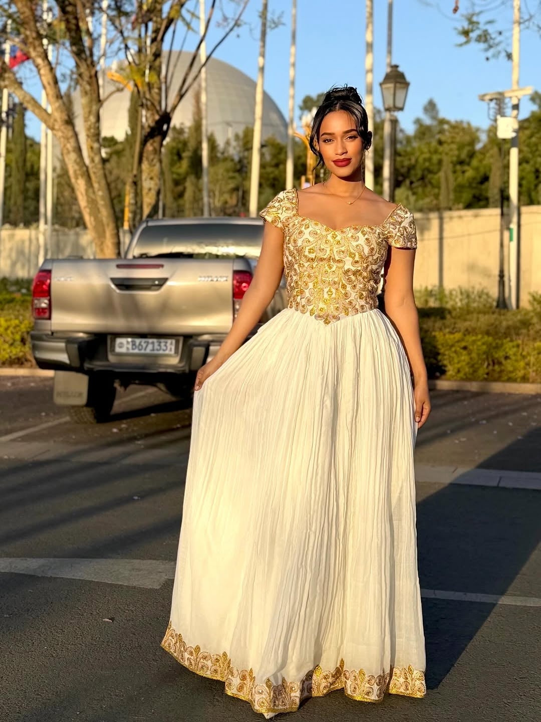 Woman in a gold and white Ethiopian cultural dress standing on a street with a car and trees in the background.