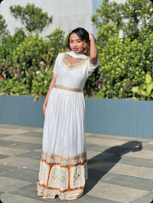 A woman wearing a white Ethiopian clothing with gold embroidery and a patterned hem, standing outdoors on a paved walkway with green foliage and a blue wall in the background.