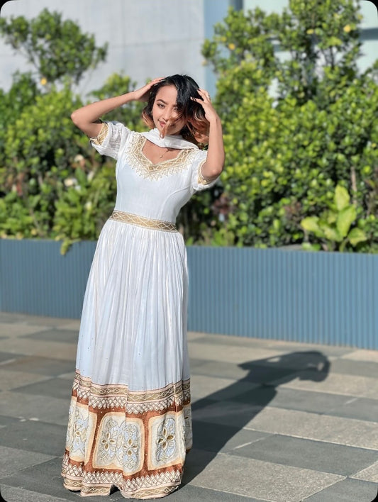 A woman wearing a long white Ethiopian clothing with gold embroidery and a patterned hem, posing outdoors on a paved walkway with green plants and a blue wall in the background.