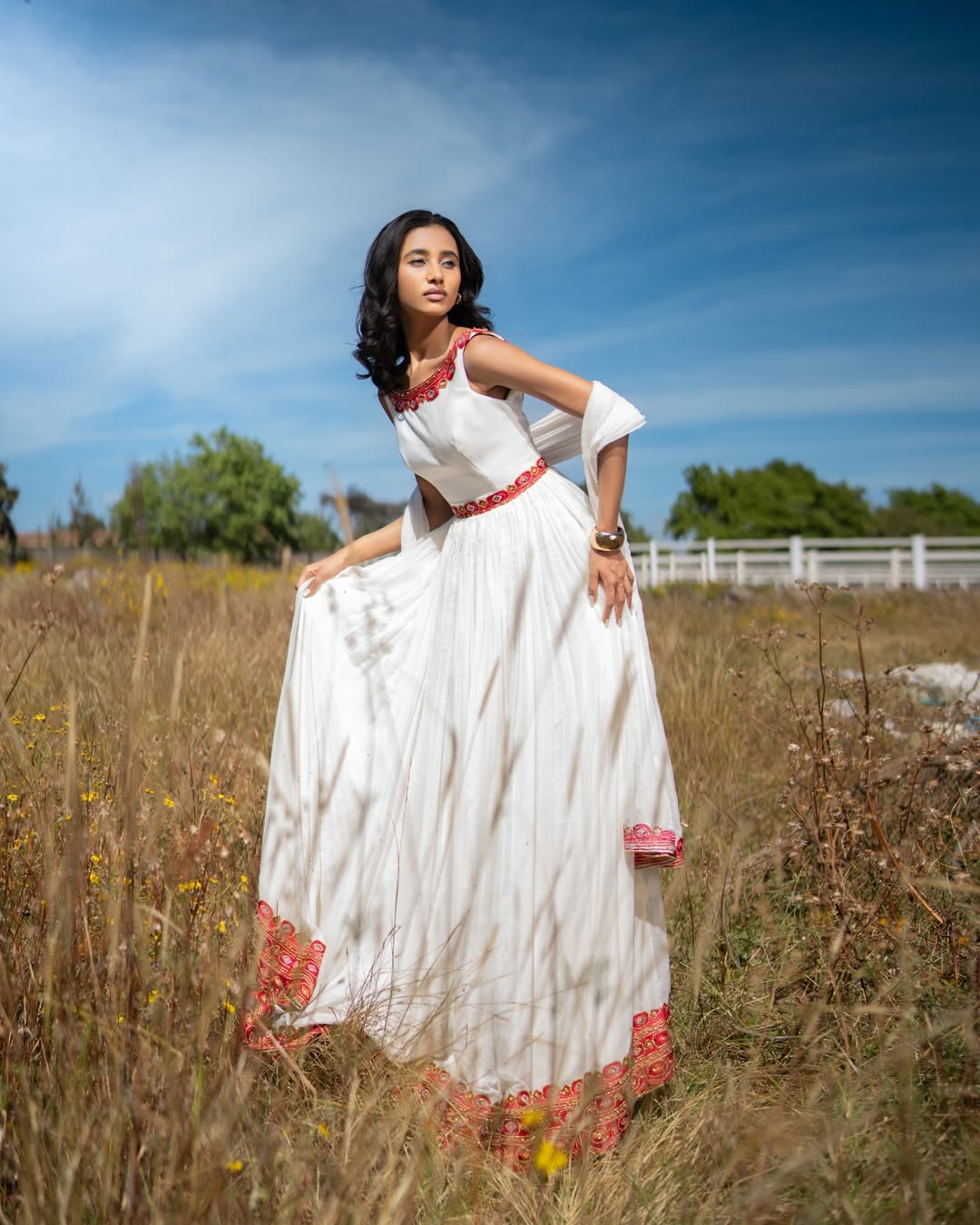 Woman in a habesha kemis with red accents standing in a field with a blue sky.