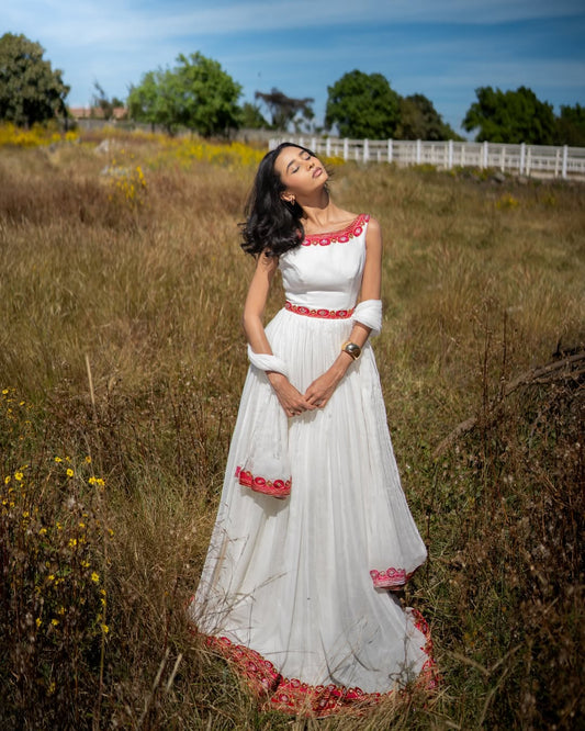 Woman in a habesha kemis with red accents standing in a field with trees and a fence in the background.