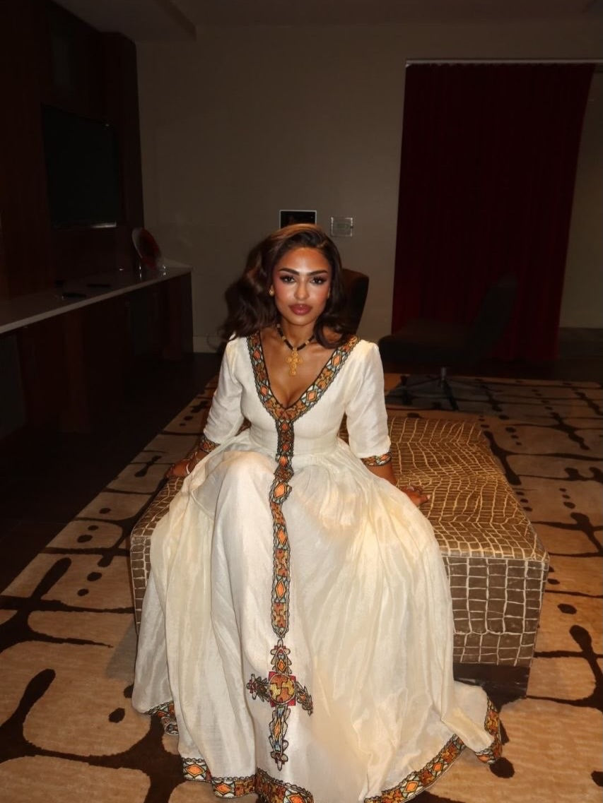 A woman wearing a white traditional Habesha Kemis with colorful embroidered details sits on a patterned bench in an indoor setting.