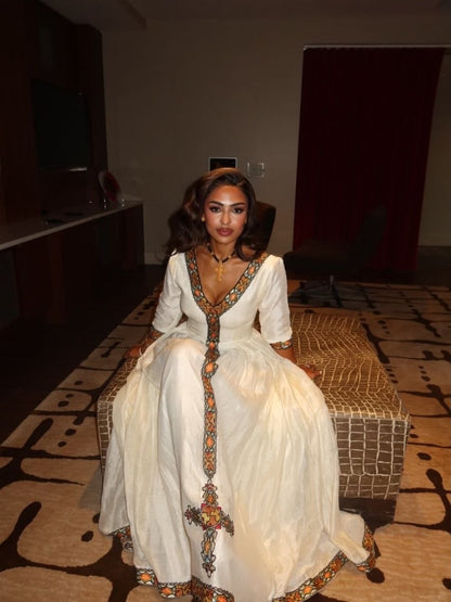 A woman wearing a white traditional Habesha Kemis with colorful embroidered details sits on a patterned bench in an indoor setting.