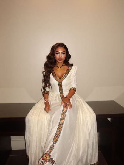A woman wearing a white traditional Habesha Kemis with colorful embroidered details, sitting on a dark bench against a plain beige wall.